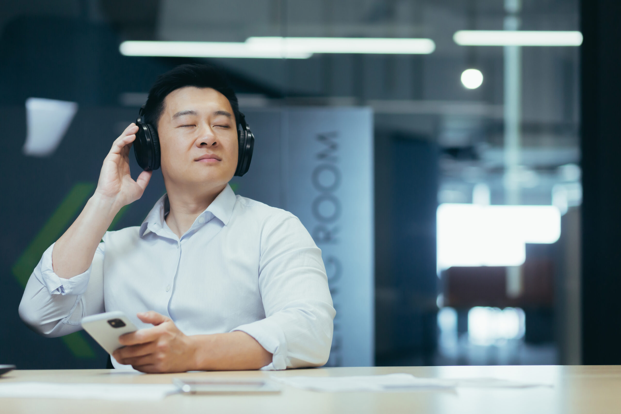 Asian boss business owner relaxing in office, listening relaxing and relaxing music, man sitting at desk, using big headphones and online listening app on phone