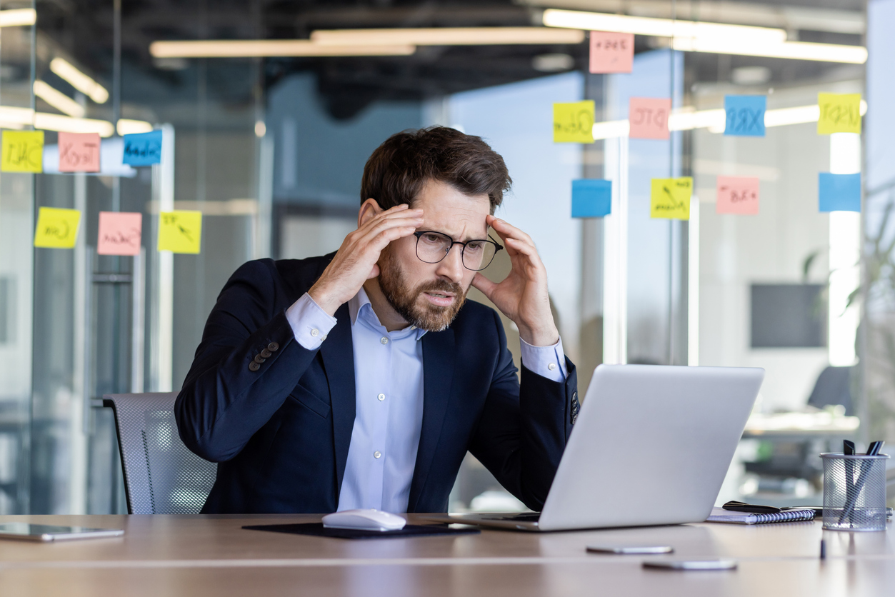 Businessman feeling stressed while working on laptop in modern office. Office space filled with colorful sticky notes. Concept of stress, productivity, and business challenges